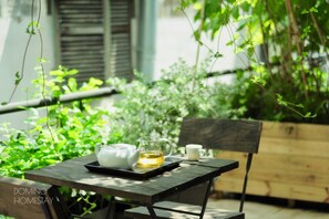 Una mesa de madera con una tetera, una jarrón y una taza, ubicada en un espacio exterior verde y exuberante con un banco de madera y una maceta.