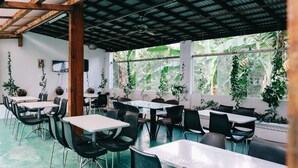 Un espace intérieur spacieux, aménagé avec des tables et des chaises, de grandes fenêtres ornées de plantes et un ventilateur de plafond.
