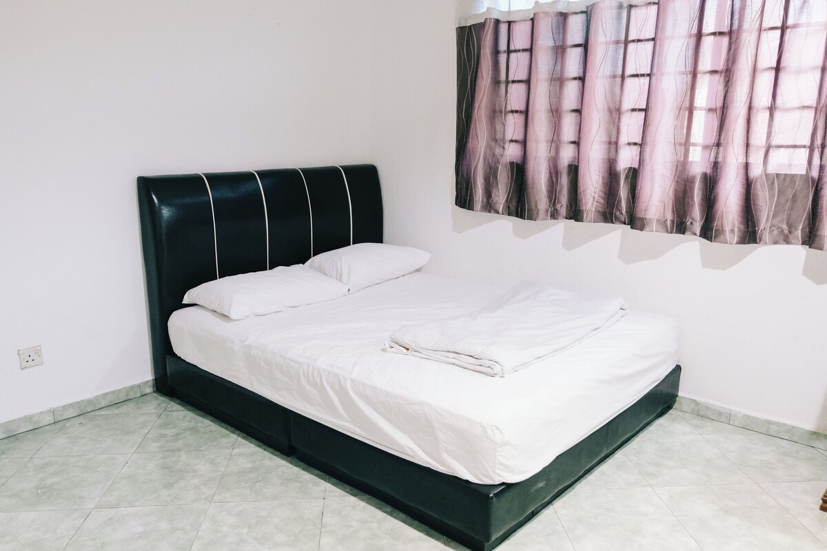 A neatly made bed in a room with a dark headboard, a plain white wall, and a window with a patterned curtain.