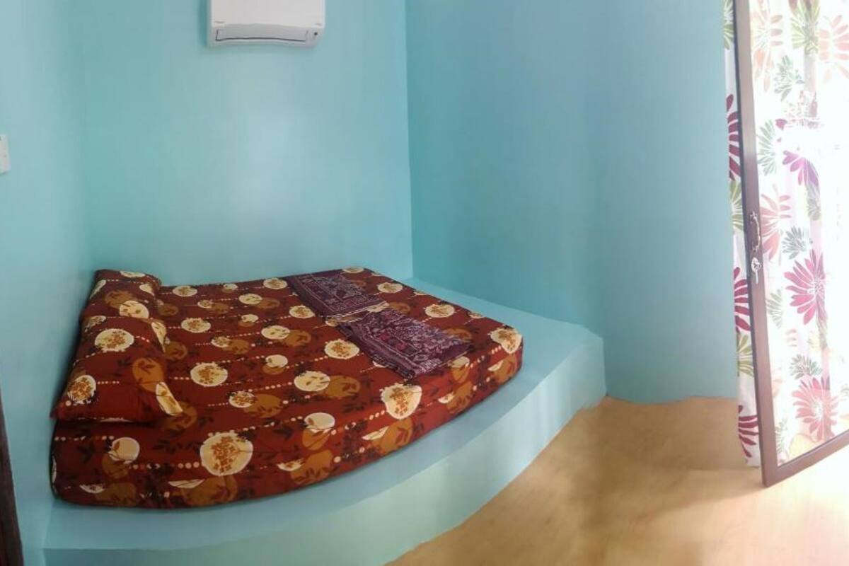 A corner room with a curved bench against a light blue wall, a small white air conditioning unit, and a window with a floral curtain.