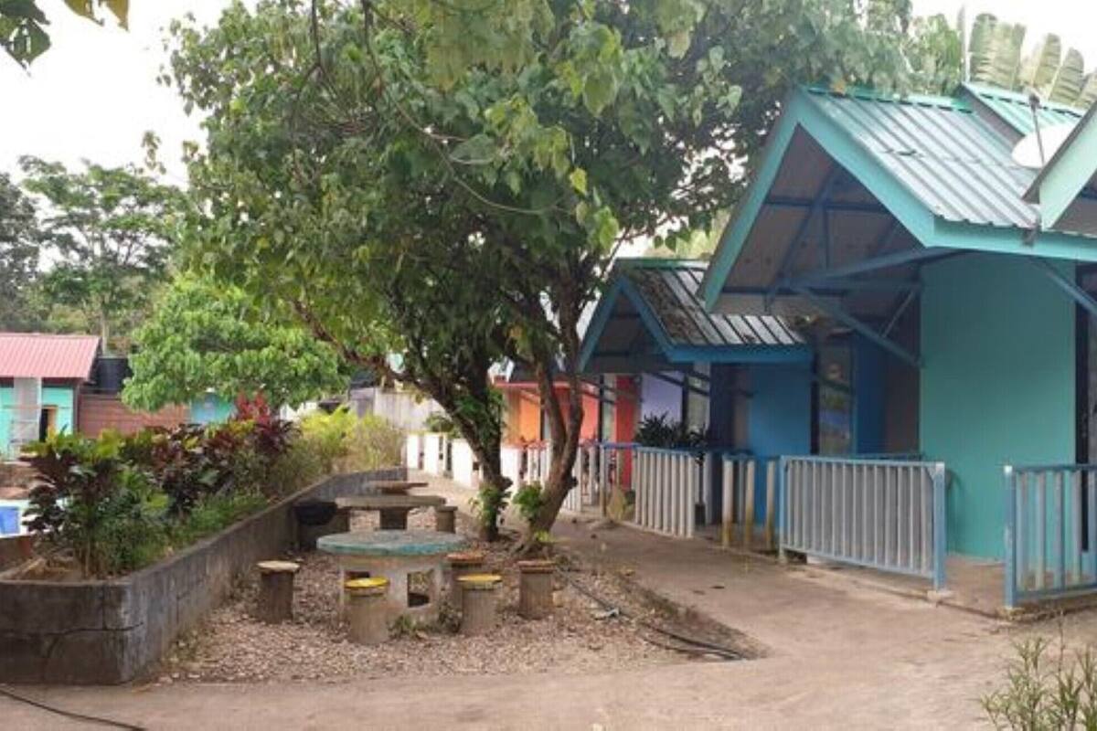 A row of blue-roofed buildings with white railings, surrounded by greenery and a gravel path.
