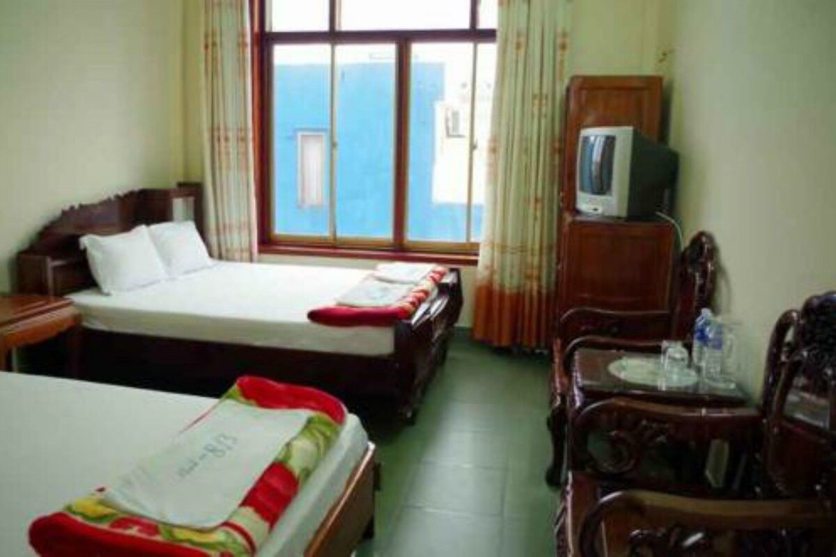A hotel room with two beds, a television, a small table with a chair, and a window with curtains.