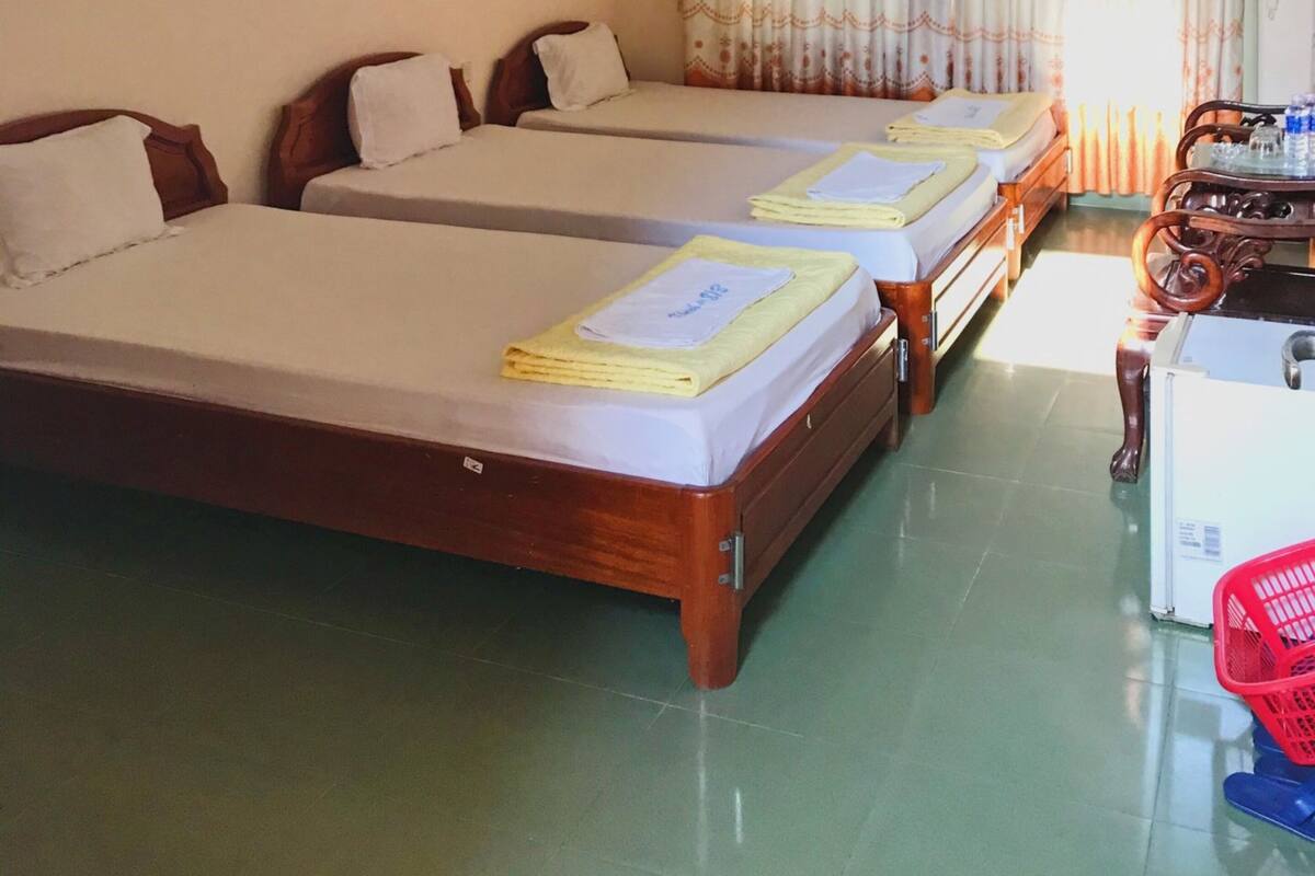 Une chambre d’hôtel avec deux lits simples, une petite table avec un ventilateur, un radiateur et un panier rouge posé sur le sol.