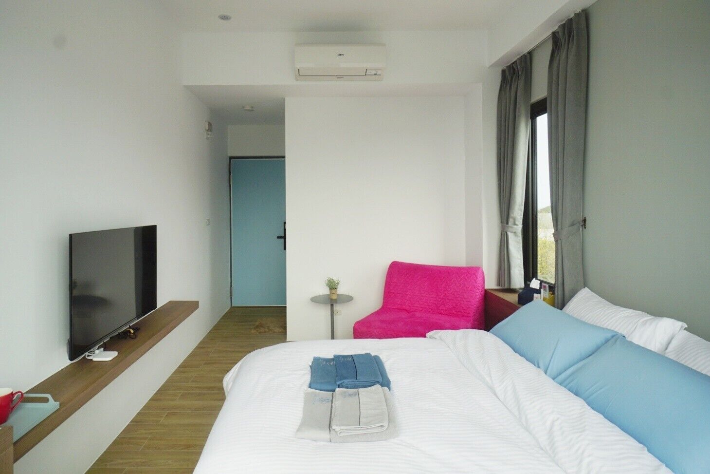 A hotel room with a bed, a TV, a small table with a plant, and a window with curtains.