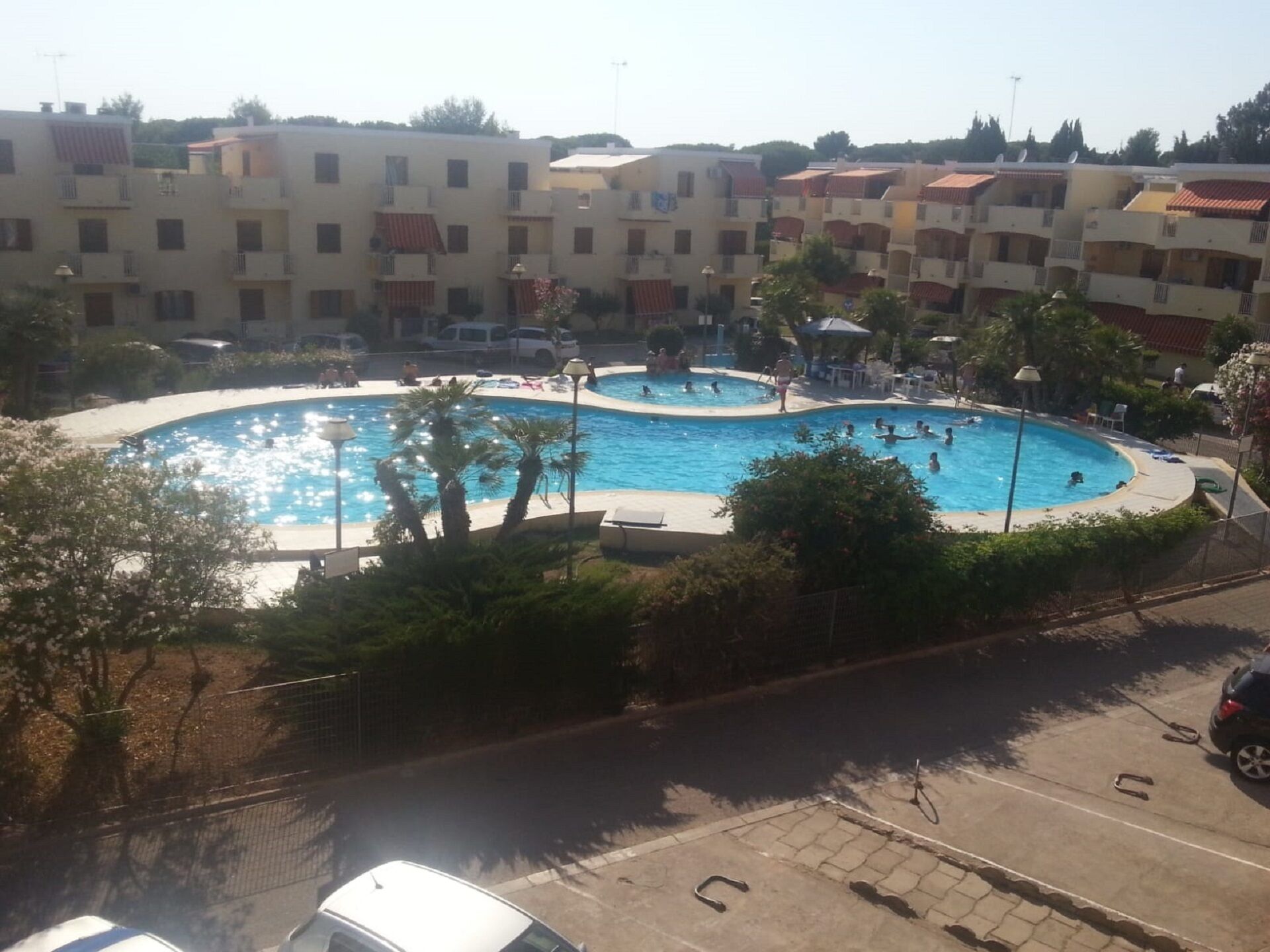 A swimming pool complex with multiple pools and surrounding greenery, set against a backdrop of residential buildings.