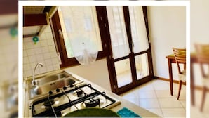 Une cuisine équipée d’une cuisinière à gaz, d’un évier et d’une fenêtre avec des rideaux. Une table à manger et des chaises se trouvent dans la pièce adjacente.