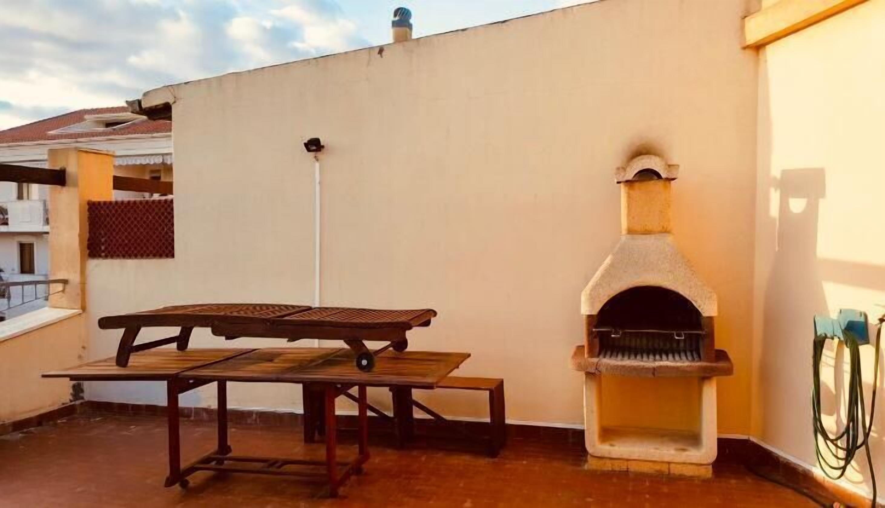 A rooftop patio with a wooden table and benches, a stone-built pizza oven, and a view of the surrounding buildings.