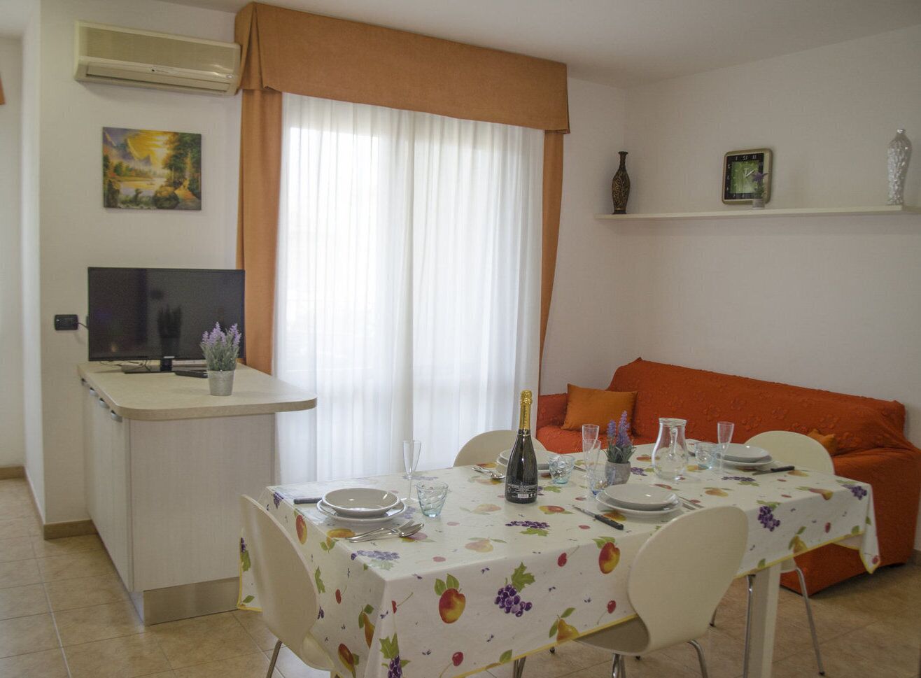A dining area with a table set for two, a television on a stand, and a sofa in the background.