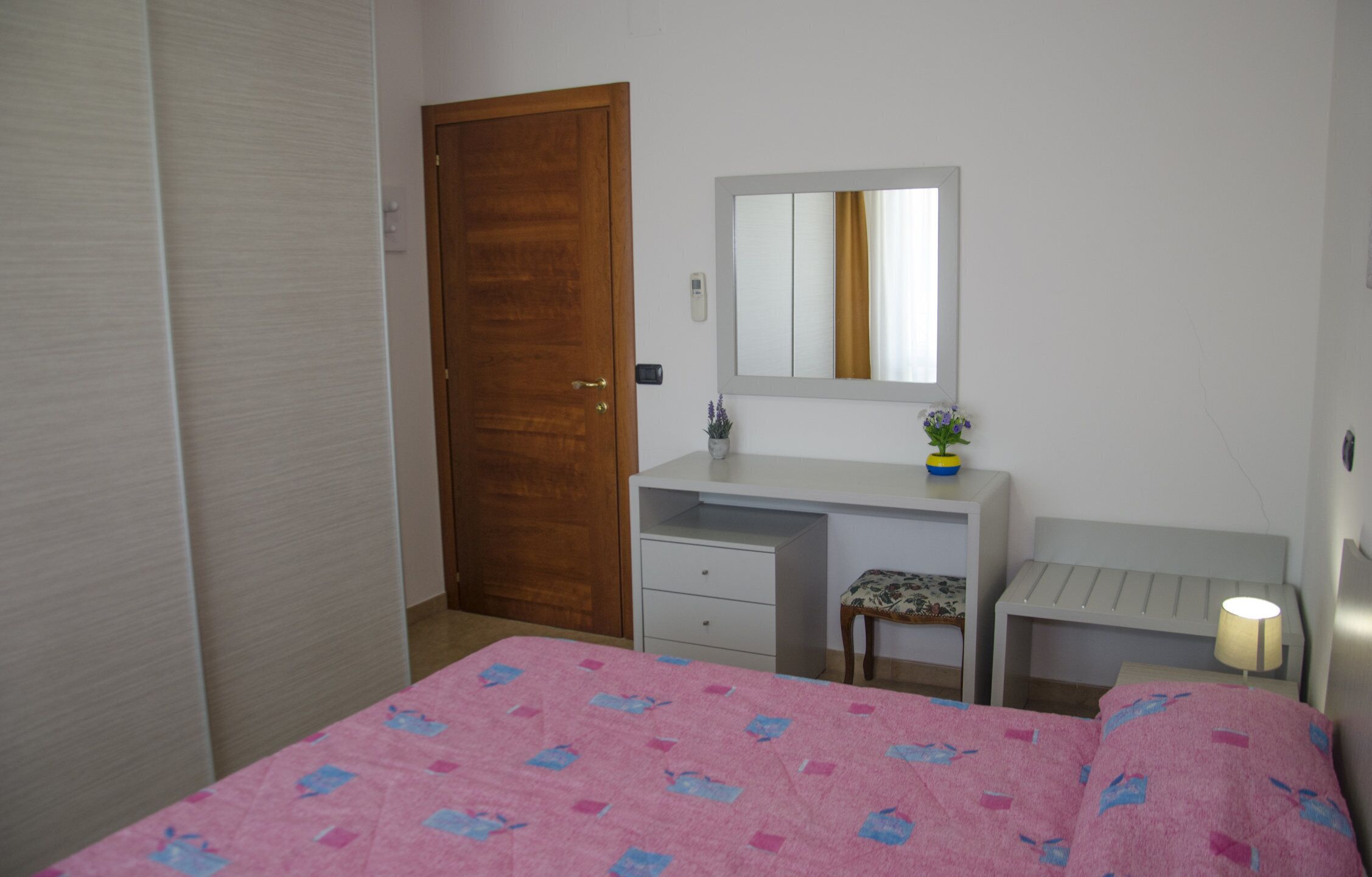 Un dormitorio con puerta de madera, cómoda blanca, mesita de noche con lámpara y cama con manta rosa.