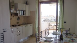 Una cocina con estufa a gas, lavamanos y gabinetes. Se ve una mesa de comedor con sillas y un balcón a través de la puerta.