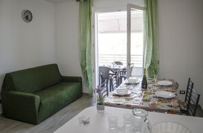 Zona pranzo con tavolo apparecchiato per due persone, un divano verde e una sveglia appesa al muro.