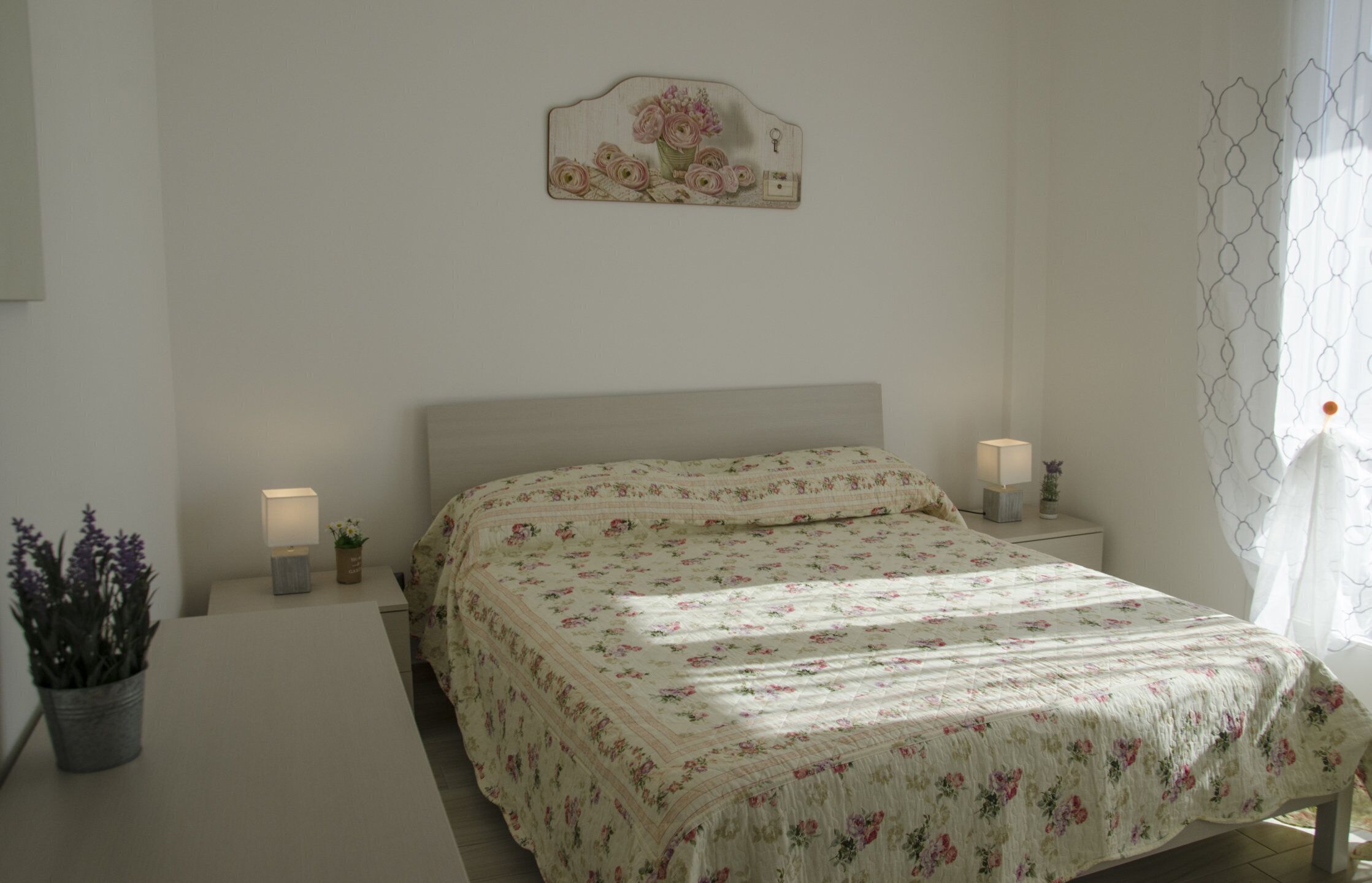 A bedroom with a floral bedspread, bedside lamps, a small potted plant, and a wall-mounted decoration.