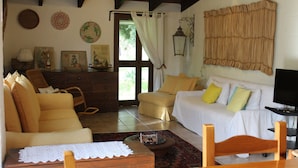 Uma sala de estar acolhedora com um sofá, uma mesa de centro de madeira e uma televisão. Há uma porta que dá para uma varanda com vista para o verde.