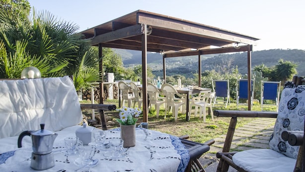An outdoor dining area with a covered pergola, tables, chairs, and a view of the surrounding landscape. - Casa Valverde (Alghero)