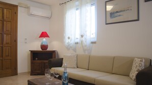 A living room with a sofa, a wooden table with wine glasses, a red lamp, and a window with sheer curtains. - Casa Valverde (Alghero)