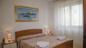 A neatly made bed with floral bedding, a nightstand with a lamp, and a painting of a coastal scene on the wall.