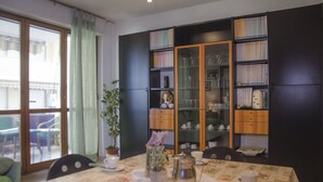 A dining area with a table set for two, a cabinet with glassware, and a view through a window.