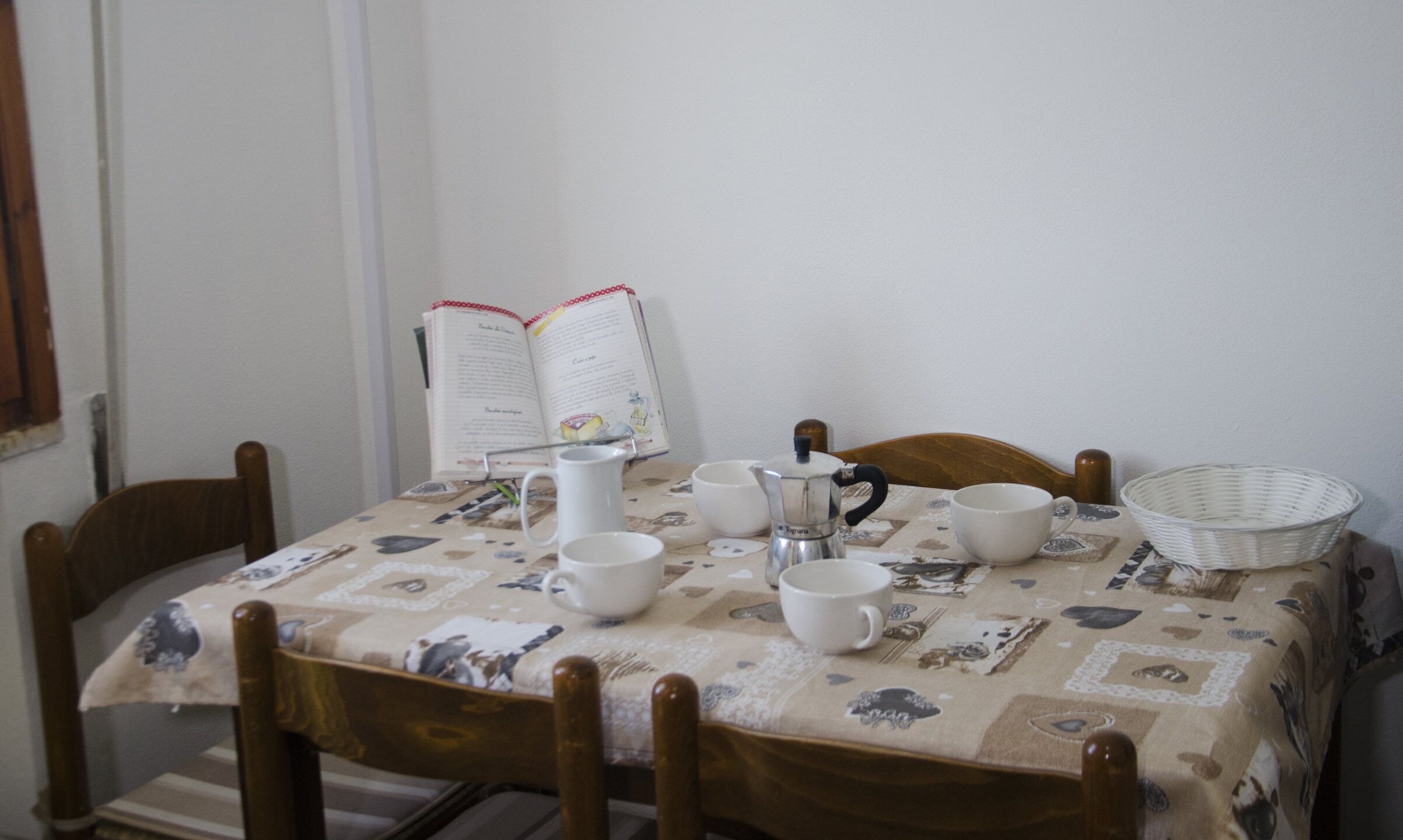 Uma mesa de jantar com toalha de mesa florida, um livro e vários copos, além de uma cafeteira.