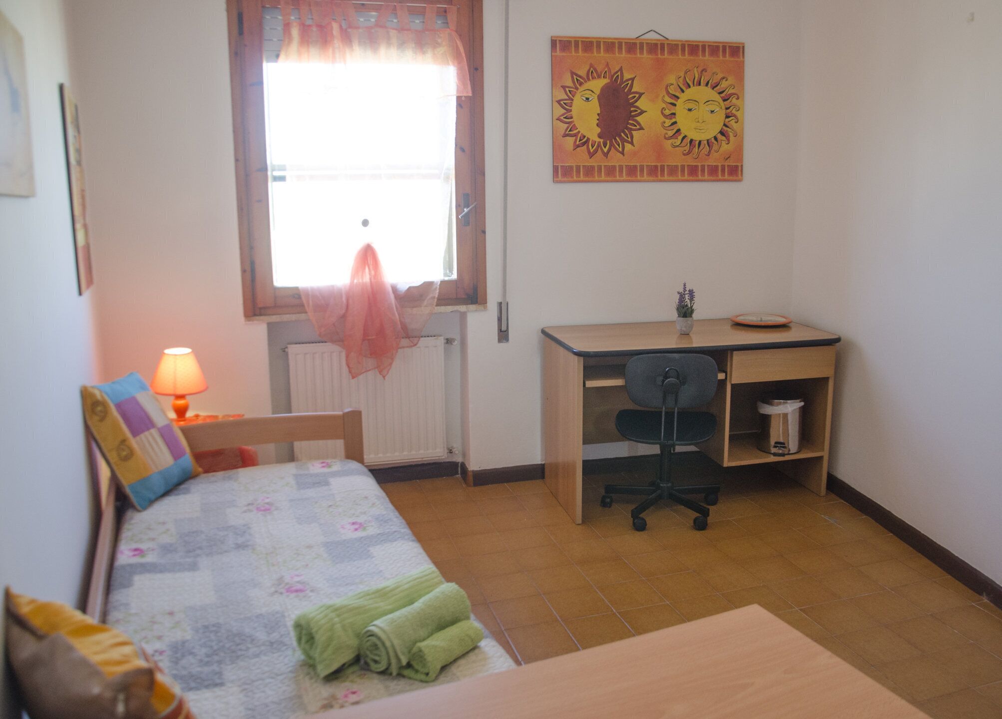Una habitación pequeña y ordenada con una cama, un escritorio, una silla, una lámpara y una ventana con cortinas.