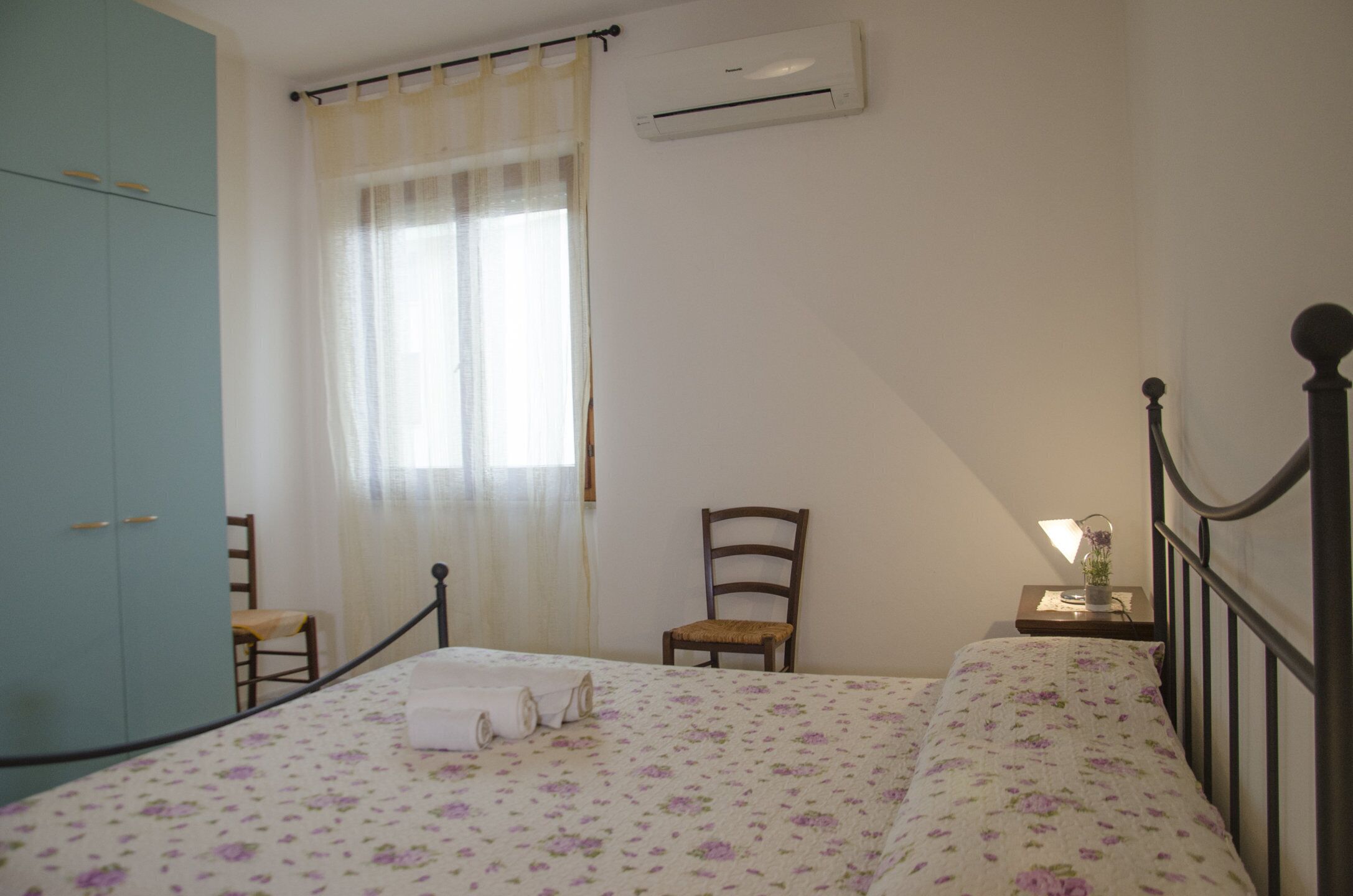A bedroom with a bed, two chairs, a wardrobe, and an air conditioning unit.