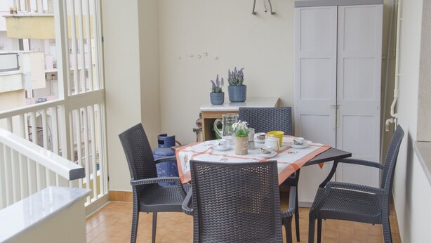 A balcony with a table set for two, featuring wicker chairs, a tablecloth, and potted plants. - Casa Lampuga (Alghero)