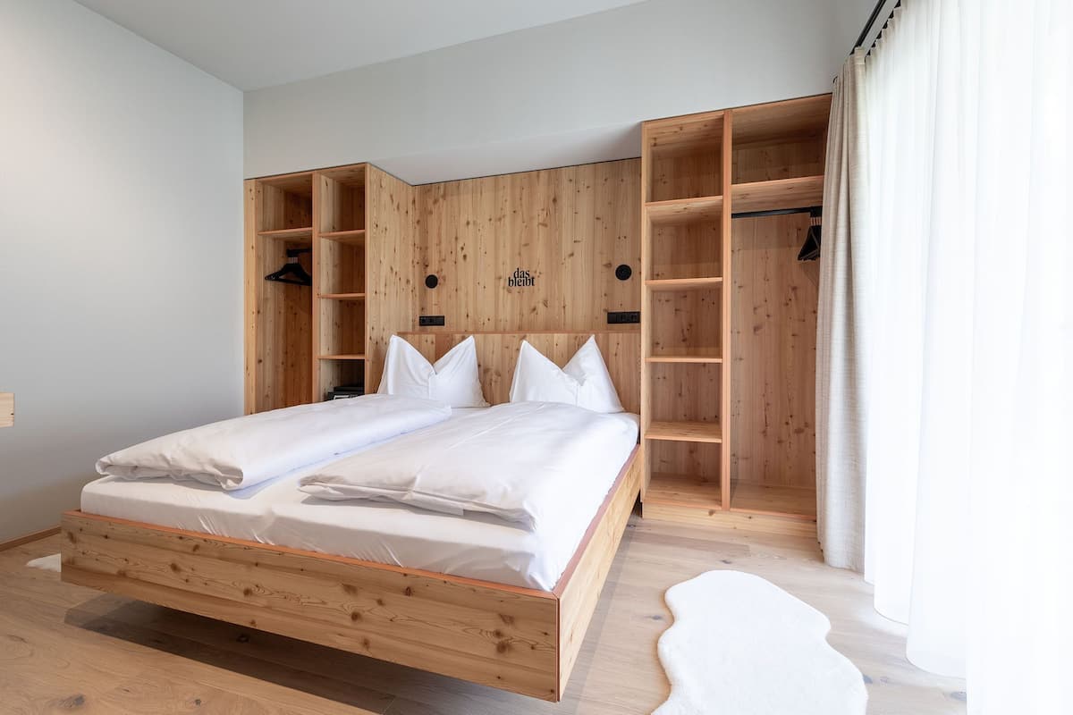 A double bed with wooden headboard and storage shelves, a white rug, and a window with curtains.