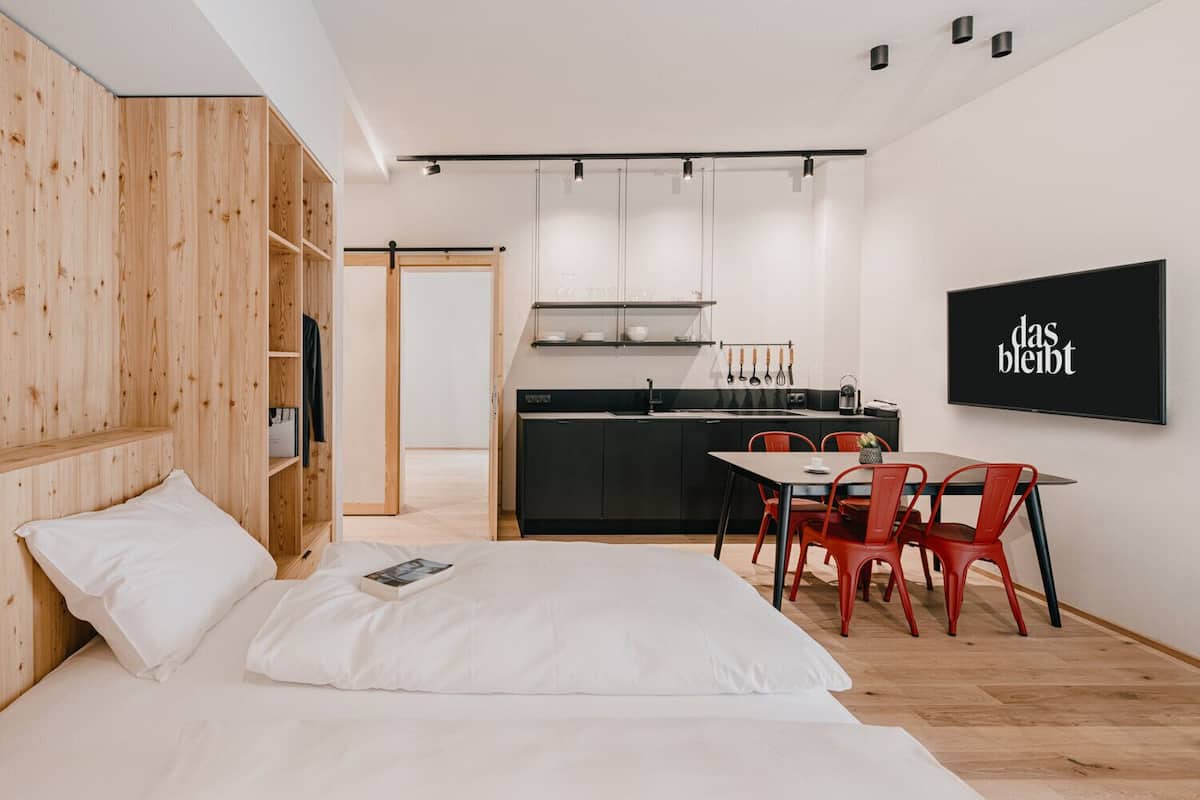 A modern bedroom with a bed, a small dining area with red chairs, and a kitchen in the background.