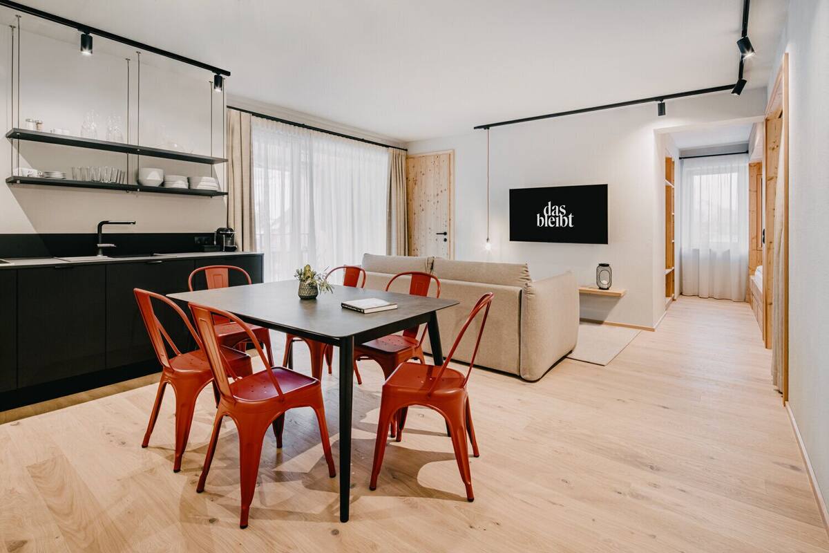 A modern living space with a dining area, kitchen, and a TV displaying 'das bleibt'.