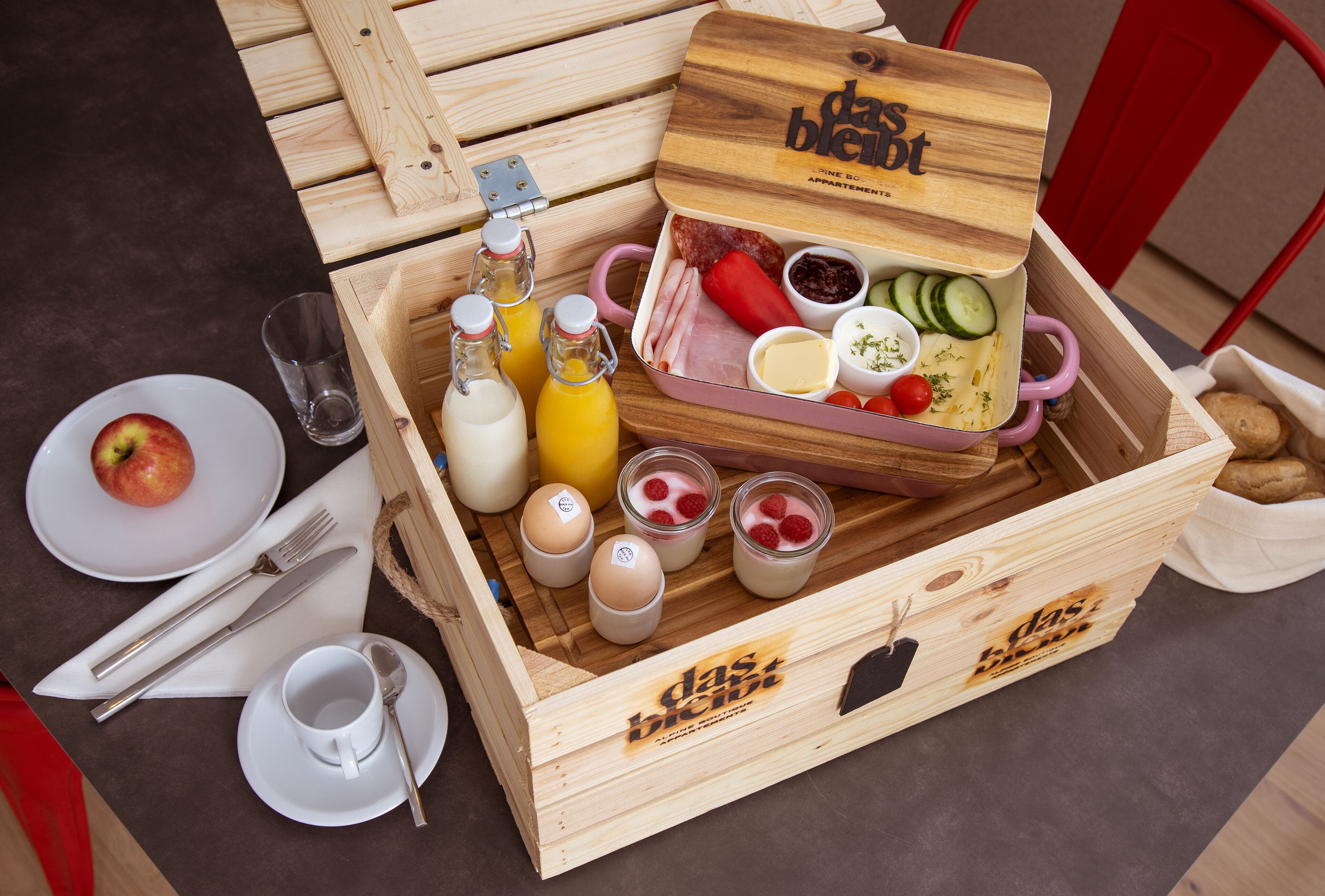Una caja de madera con tapa etiquetada “das blend” que contiene alimentos para el desayuno, como huevos, queso y fruta.
