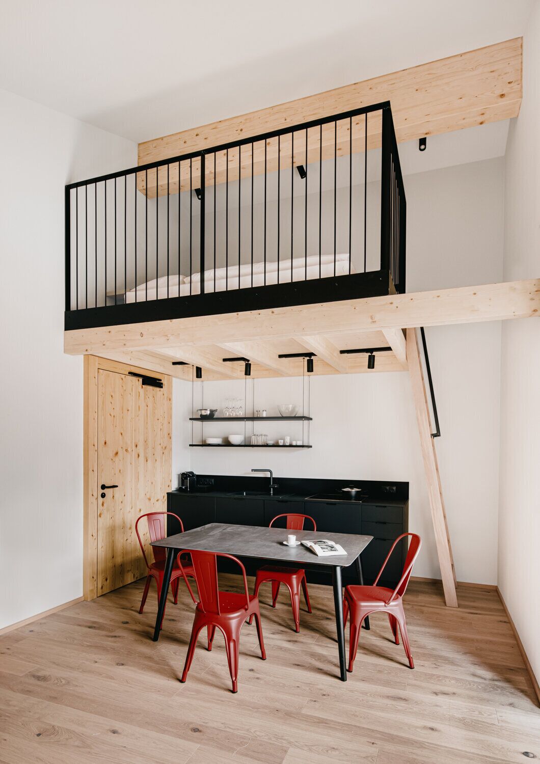 A compact kitchen with a dining area, featuring a wooden ceiling, black railings, and red chairs.