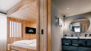 A modern bedroom with a wooden headboard, a bed, a TV, and a bathroom with a mirror and sink. - das bleibt (Schladming)