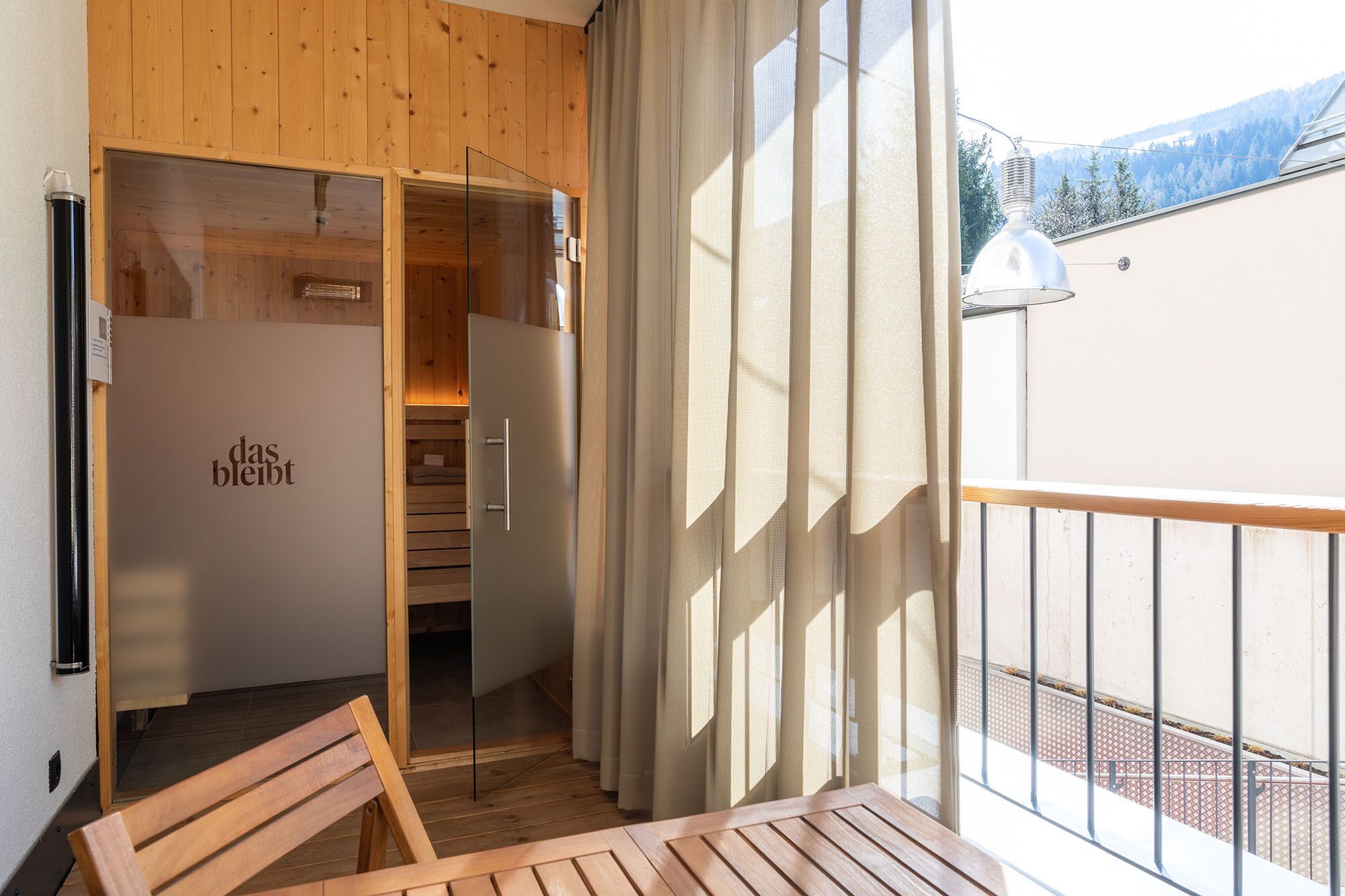 A wooden interior with a sliding door leading to a sauna room, a wooden bench, and a balcony with a view of the mountains.