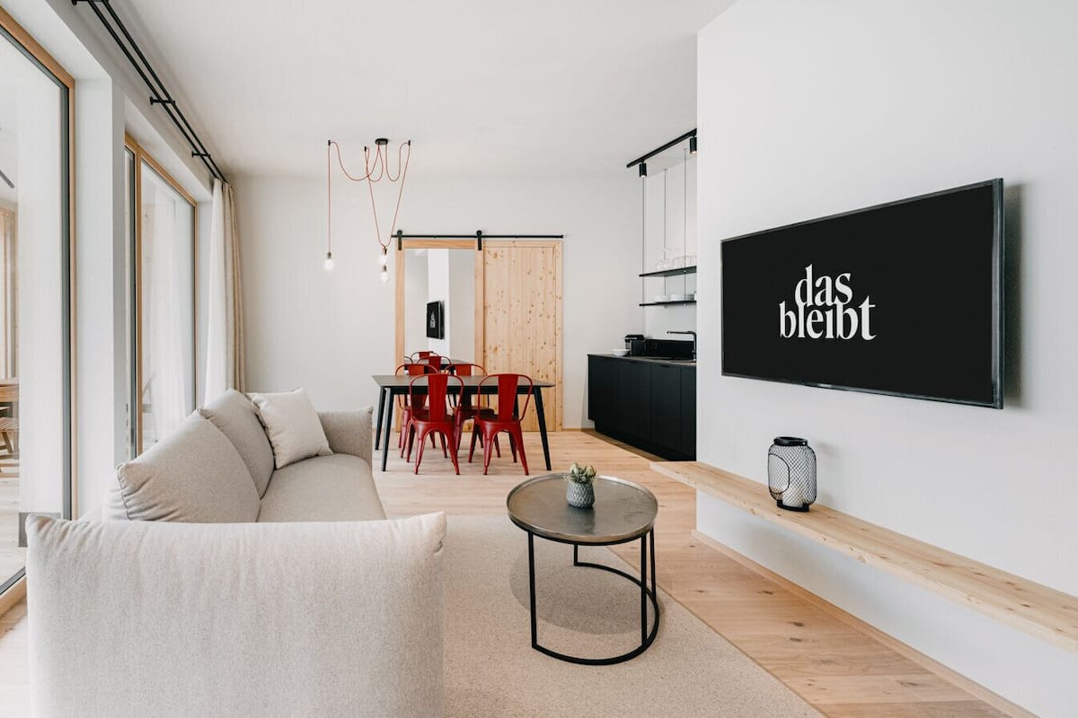A modern living room with a sofa, a dining table with chairs, a TV mounted on the wall, and a small table with a vase.