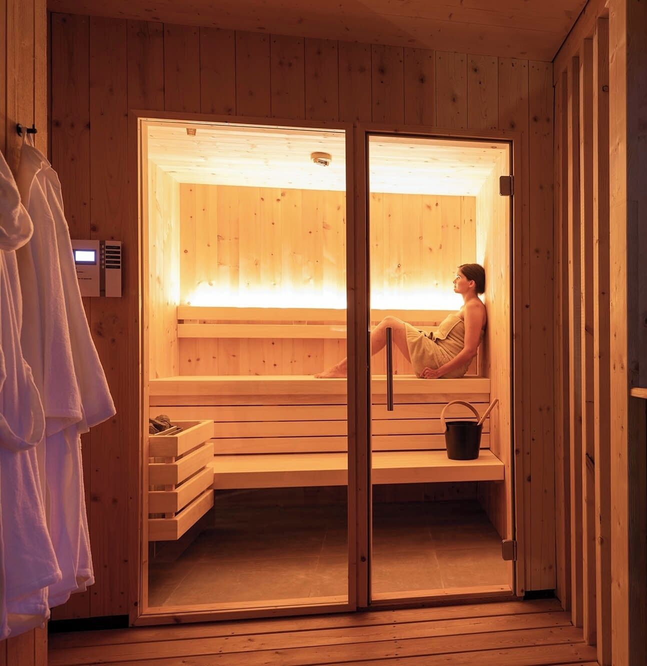 Una sauna con banco, balde, paredes de madera y panel de control visible.
