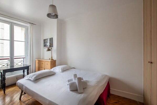 A neatly made bed with white linens and towels, a wooden nightstand with a lamp, a small wooden dresser, and a window with a view of buildings. - Spacious flat in Batignolles quarter (Paris)
