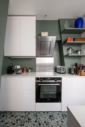 A compact kitchen with white cabinets, a stainless steel range hood, a built-in oven, and a countertop with various appliances and utensils. - Spacious flat in Batignolles quarter (Paris)