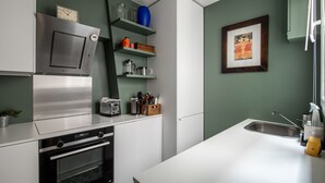 A modern kitchen with a built-in oven, microwave, and stainless steel range hood. There are shelves with kitchen items and a framed picture on the wall. - Spacious flat in Batignolles quarter (Paris)
