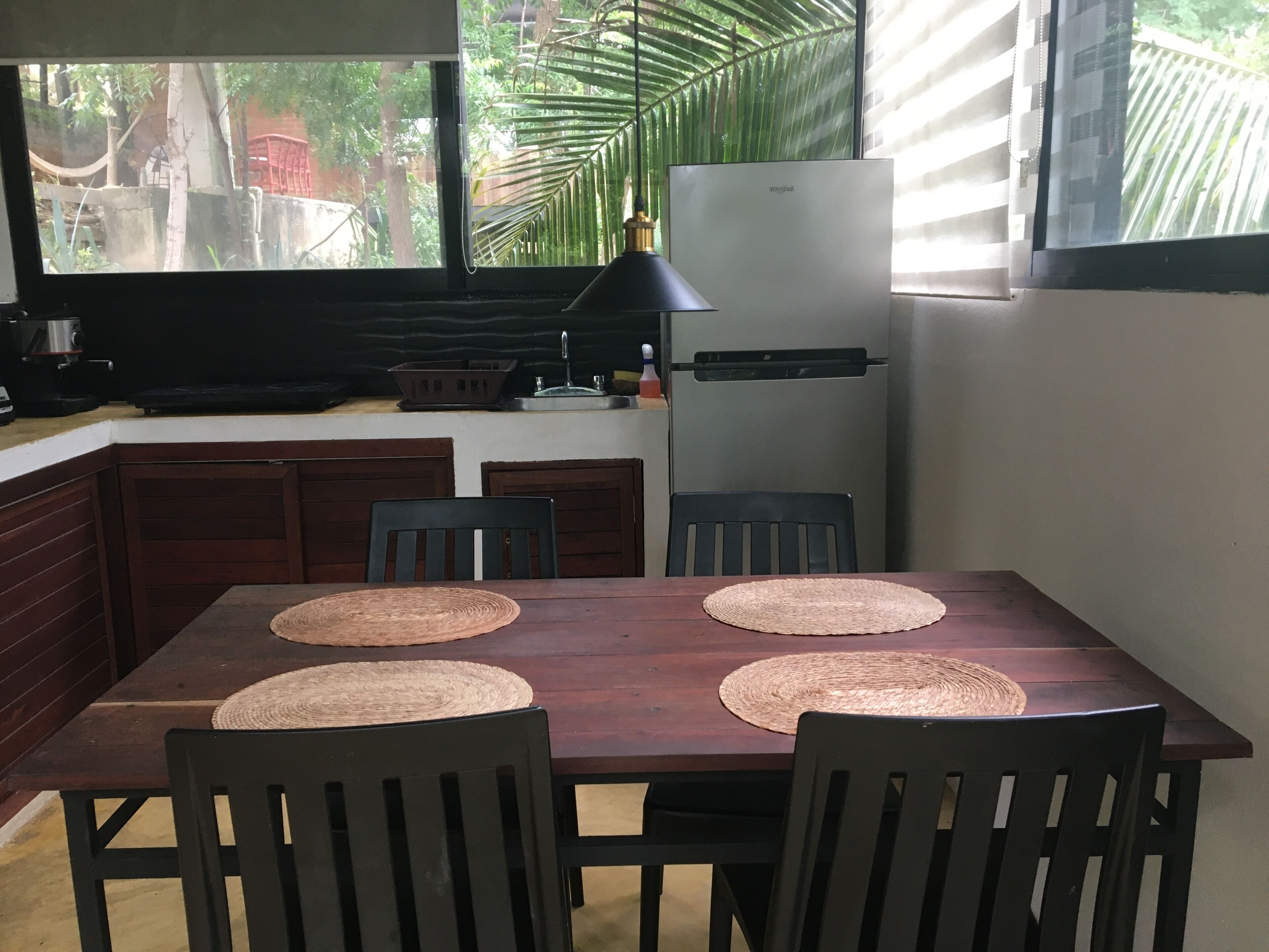 A kitchen with a dining table, chairs, a refrigerator, and a window with a view of greenery.