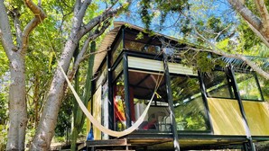 Une cabane dans les arbres dotée d’une structure vitrée, d’une terrasse en bois et d’un hamac.