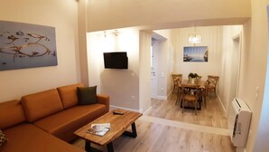 Una sala de estar con sofá, mesa de centro y televisor montado en la pared. Hay un comedor con mesa y sillas, y un cuadro en la pared.