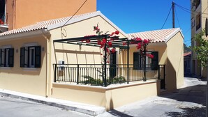 Un edificio pequeño de una sola planta con balcón, techo de tejas y un jardín con flores rojas.