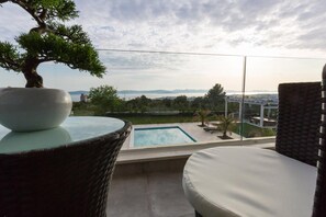 A balcony with a view of a swimming pool, a bonsai tree, and wicker furniture.