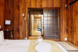 Una habitación de estilo japonés tradicional con paredes de madera, suelo de estera de tatami, una cama con ropa de cama blanca y una puerta corredera de shoji.