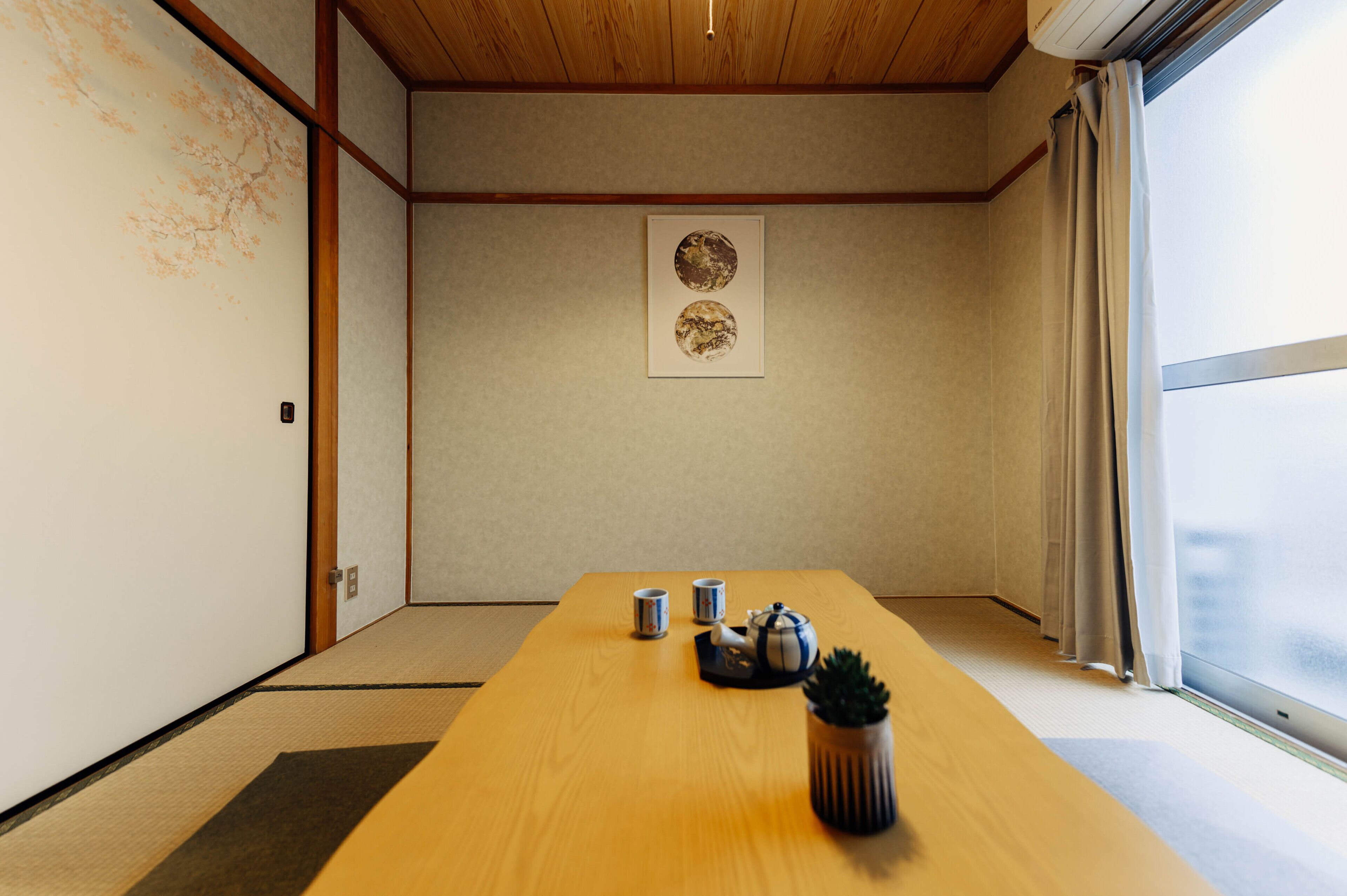 A room with tatami flooring, a wooden table with a teapot and cups, a framed picture on the wall, and a window with curtains.