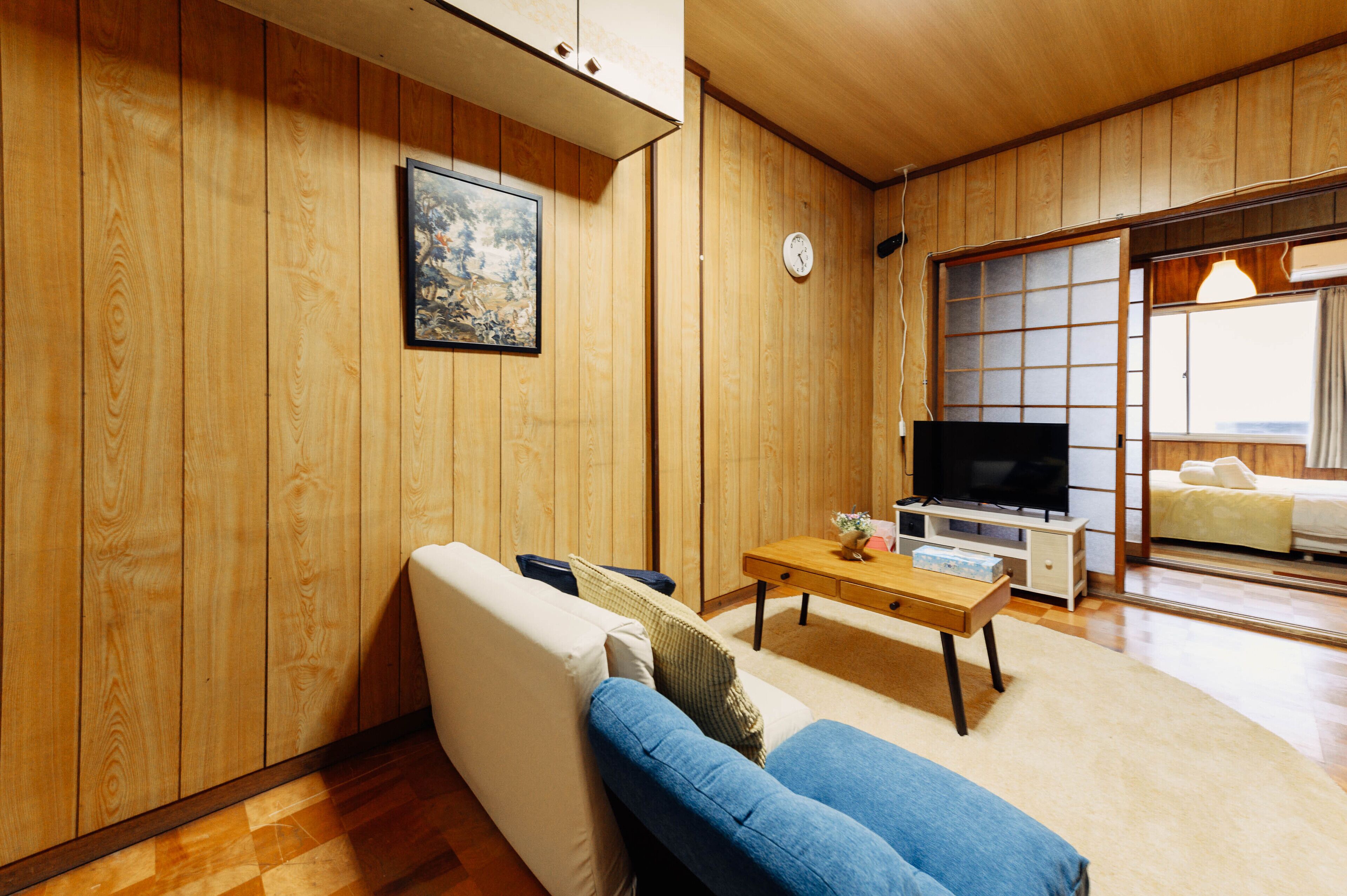 Un salon de style japonais traditionnel, avec des murs en bois, un canapé, un téléviseur et une porte coulissante donnant sur une autre pièce.