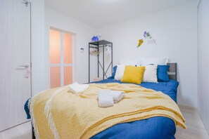 A neatly made bed with a yellow blanket and pillows, a white door, and a small shelf with items on it. - NICE HOUSE (Osaka)