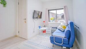 A small, bright room with a blue sofa, a TV mounted on the wall, a window with curtains, and a small table with a vase of flowers. - NICE HOUSE (Osaka)