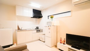 A modern kitchen with white cabinets, a microwave, and a stove. There is a flat-screen TV on a stand and a small table with decorative items. - Doraemon's House (Osaka)