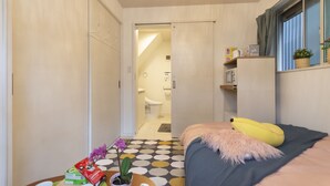 A compact bedroom with a bed, a bedside table with a banana and a plant, a window with curtains, and a view of the bathroom. - MARE HOUSE-1F (Tokyo)