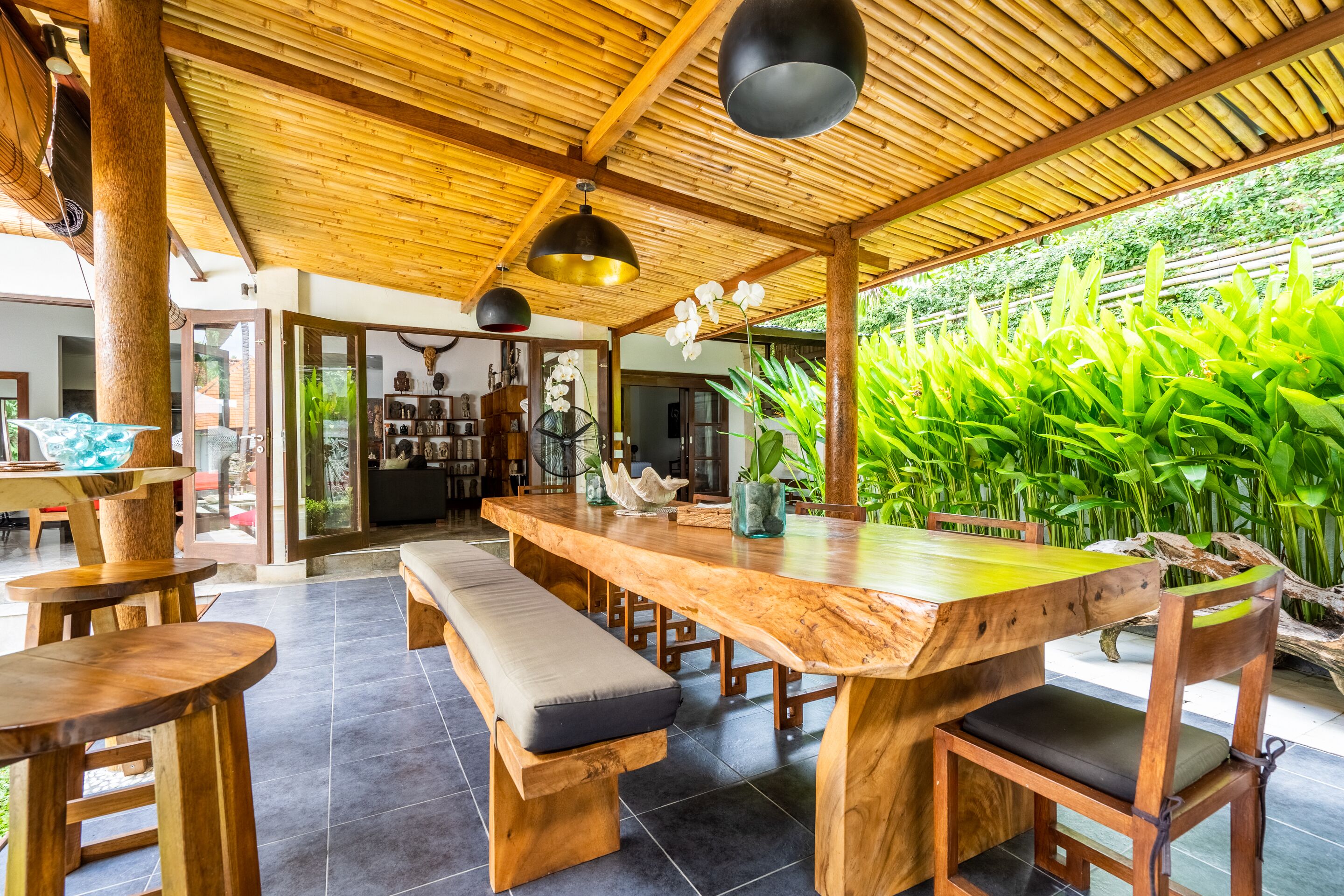 A spacious wooden dining area with a large table and benches, surrounded by lush greenery.